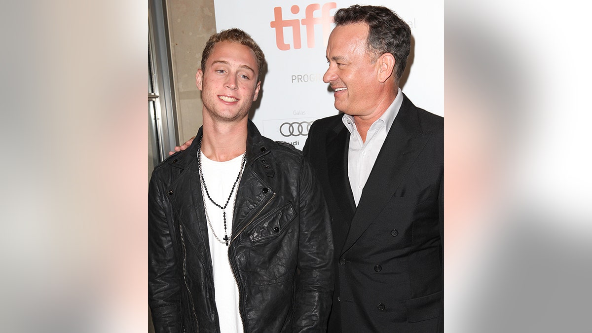 Chet Hanks in a black jacket smiles as his father Tom Hanks in a black suit puts is arm around him and looks at him lovingly