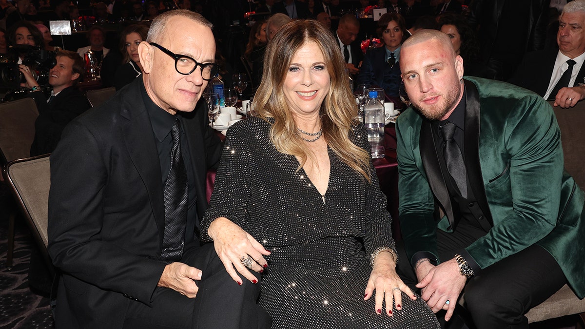 Tom Hanks in a black suit and tie sits next to wife Rita Wilson in a sparkly black dress next to son Chet Hanks in a green tuxedo jacket