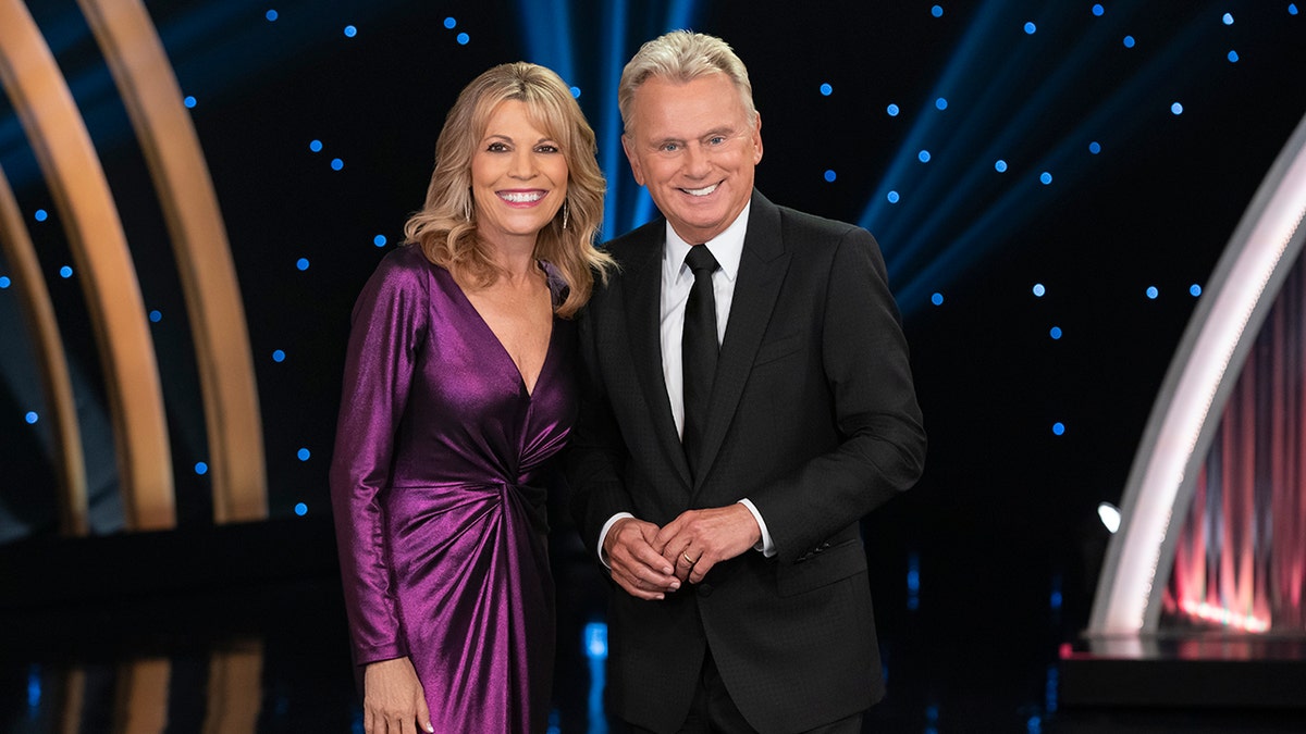 Vanna White smiles in a slinky purple dress with "Celebrity Wheel of Fortune" co-host Pat Sajak in a black suit and tie