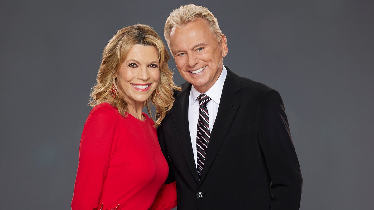 Vanna White in a red dress smiles as she poses for a photo with "Celebrity Wheel of Fortune" co-host Pat Saja in a black suit