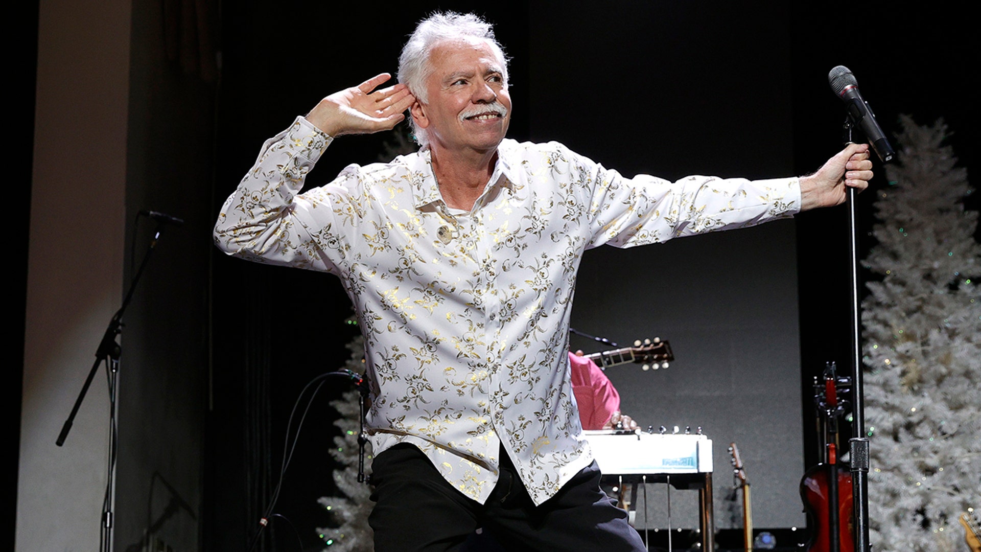 Joe Bonsall in a white patterned shirt puts his hand up to his ear while on stage in Tennessee