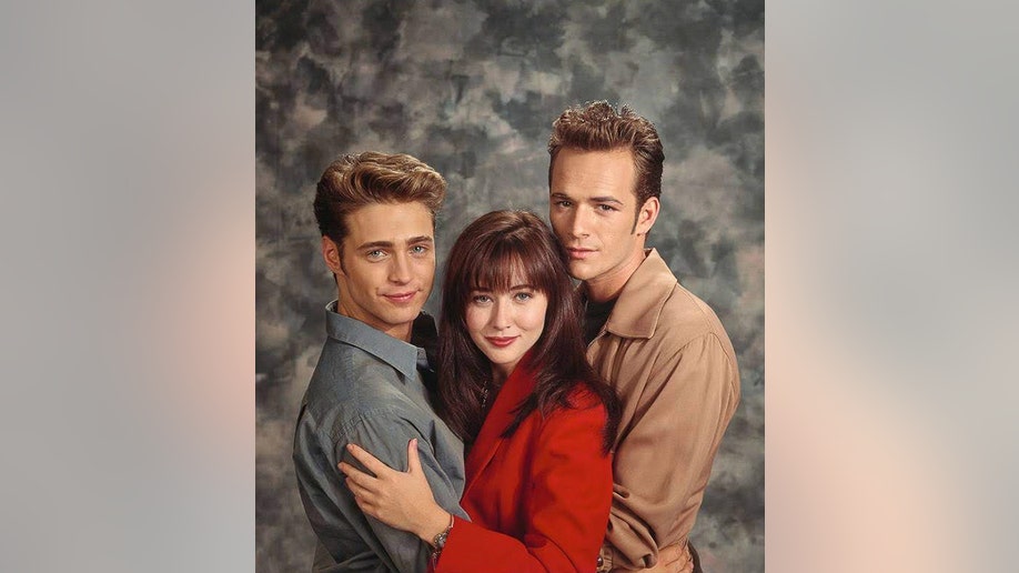 Shannen Doherty in red holds onto Jason Priestly and is hugged by Luke Perry