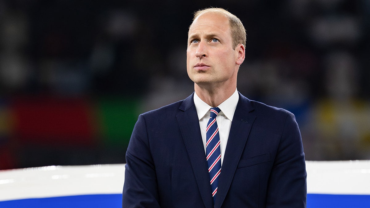 Prince William looks up wearing a navy suit and red and blue striped tie