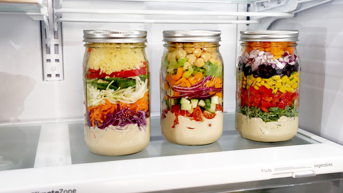 Tres tarros de cristal llenos de "ensaladas" con un aliño en el fondo. Las capas individuales son diferentes en cada tarro, pero contienen verduras, cereales y otros ingredientes.