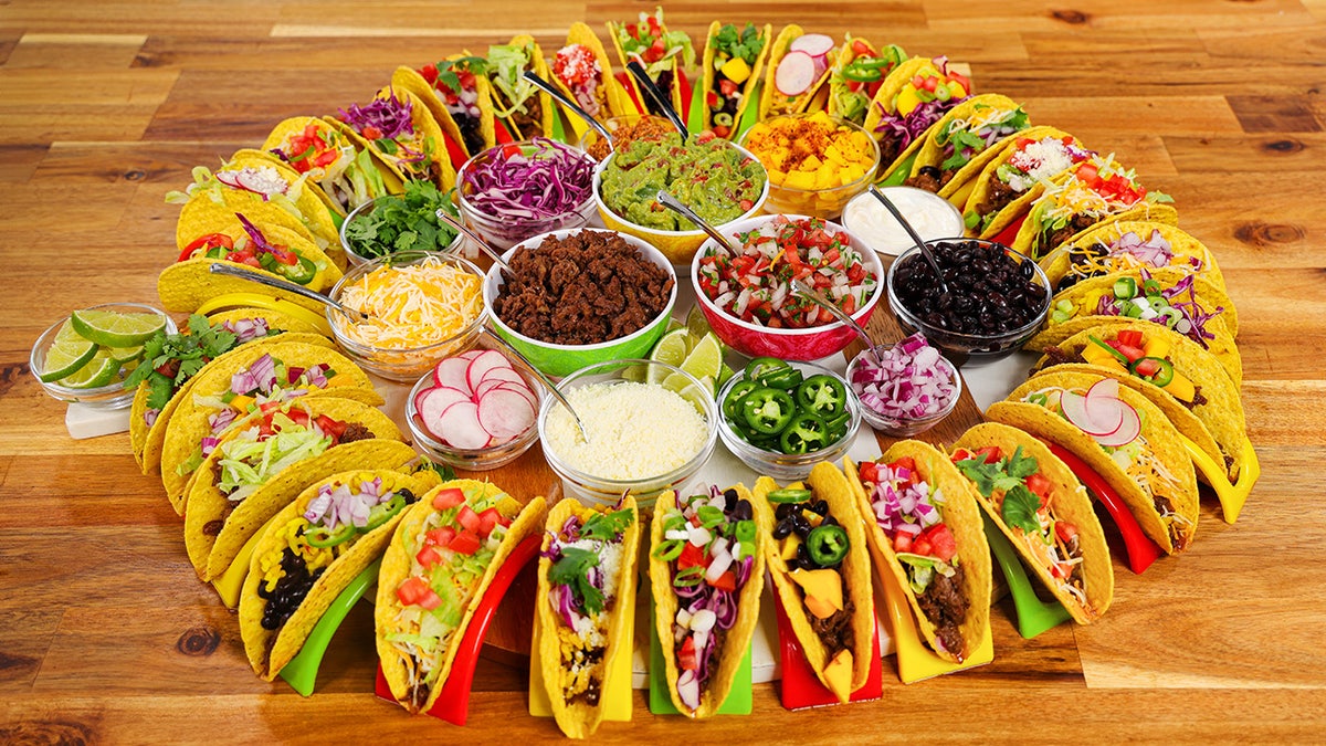 A taco board consisting of a circle of filled hard taco shells with various taco toppings in the center. 