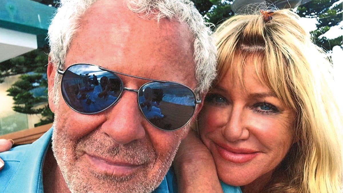 Suzanne Somers leaning on Alan Hamel who is wearing a bright blue shirt and sunglasses.