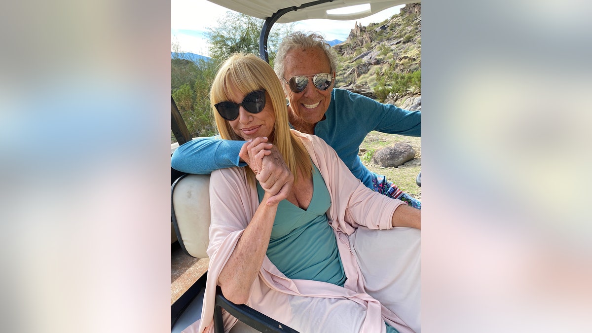 Suzanne Somers wearing a green shirt and a pink cardigan embracing Alan Hamel inside a golf cart.