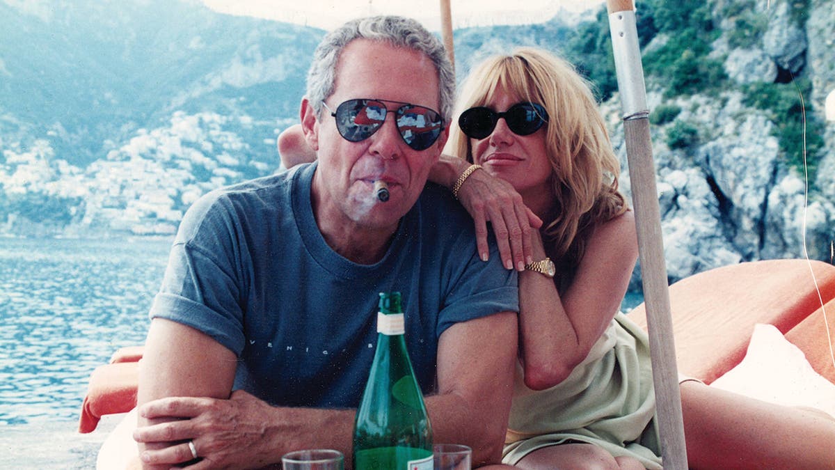 Alan Hamel smoking a cigar as Suzanne Somers leans on him outdoors beside a green glass bottle.