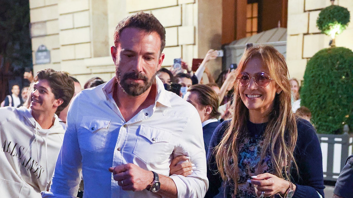 Ben Affleck parecendo sério com uma sorridente Jennifer Lopez caminhando por um espaço público