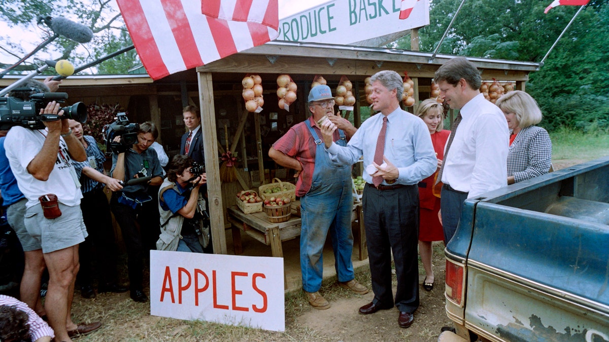 El candidato presidencial demócrata Bill Clinton y su compañero de fórmula Al Gore, flanqueados por sus esposas Tipper Gore y Hillary Clinton, hablan con Gary West en su puesto de productos agrícolas el 23 de septiembre de 1992, mientras inspeccionan algunos de los artículos.