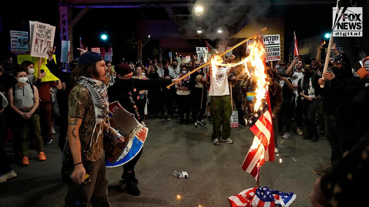 Pro-Hamas demonstranti stāv apkārt uzmundrinošiem, jo ​​Amerikas karogs deg uz zemes