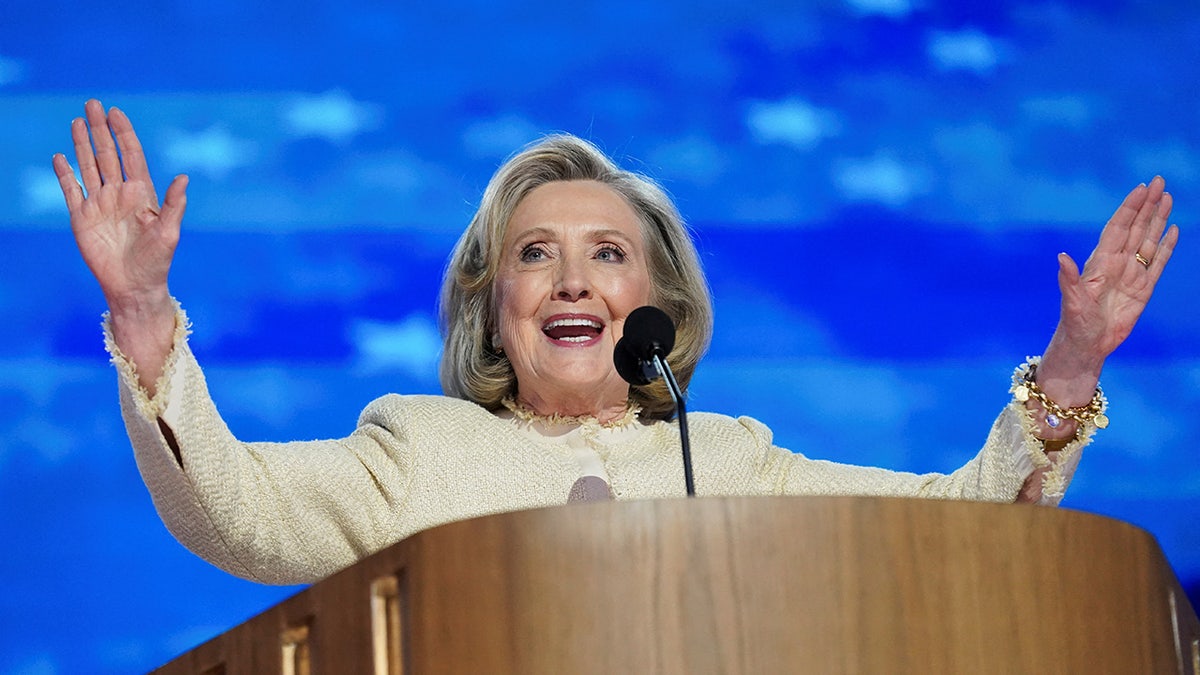 Hillary Clinton with arms spread, speaking behind podium