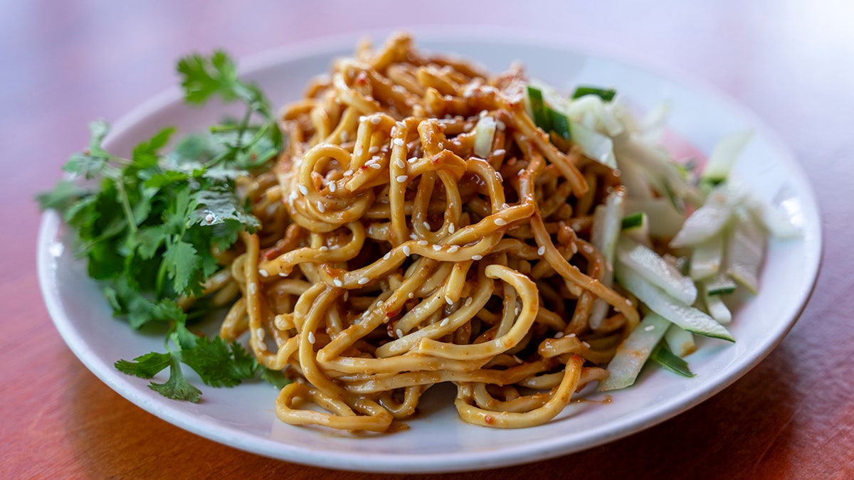 A plate of chilled egg noodles with cilantro and cucumber on the side. The noodles are tossed with chocolate sauce and sprinkled with sesame seeds.