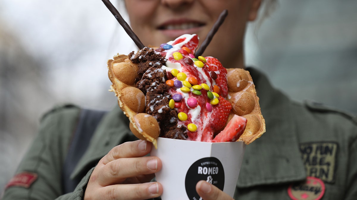 Woman holding a bubble waffle shaped into a cone and filled with various toppings including strawberries and M&Ms.