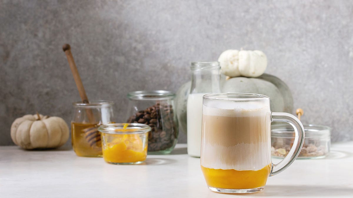 Vaso de café con leche de calabaza y especias con puré de calabaza, con espuma de leche y canela de pie con ingredientes en tarros y calabazas blancas decorativas sobre mesa de mármol blanco con pared gris de fondo.