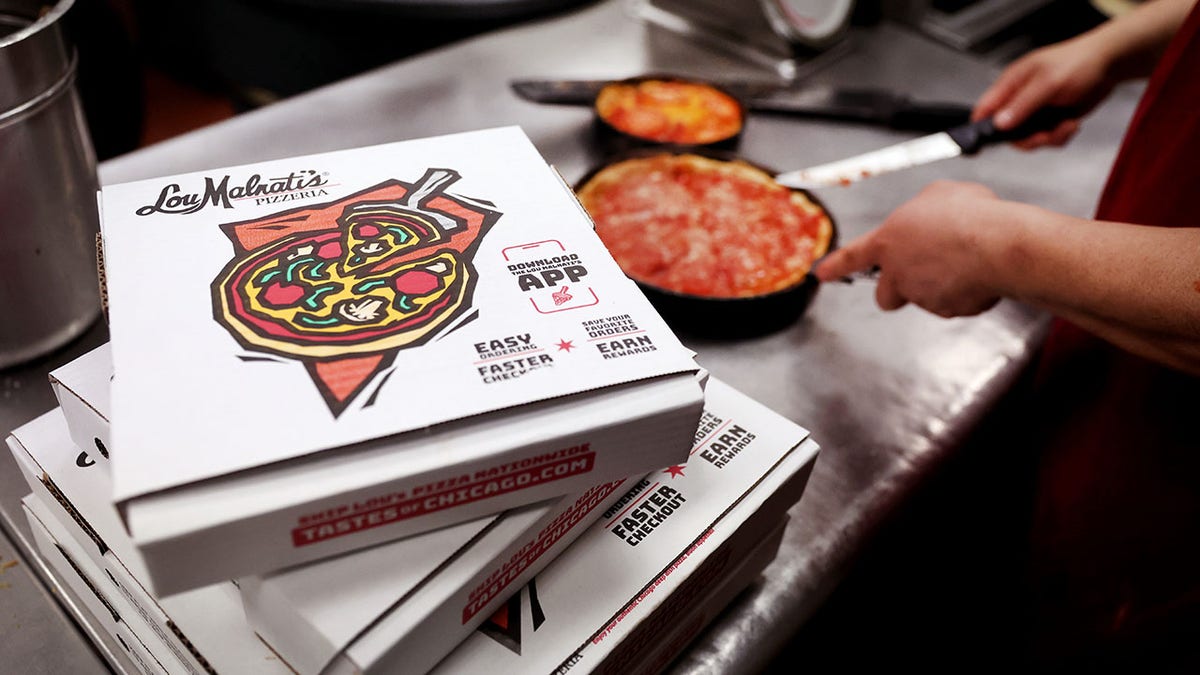 A worker making a deep-dish pizza with a pizza box on the left side of the photograph.