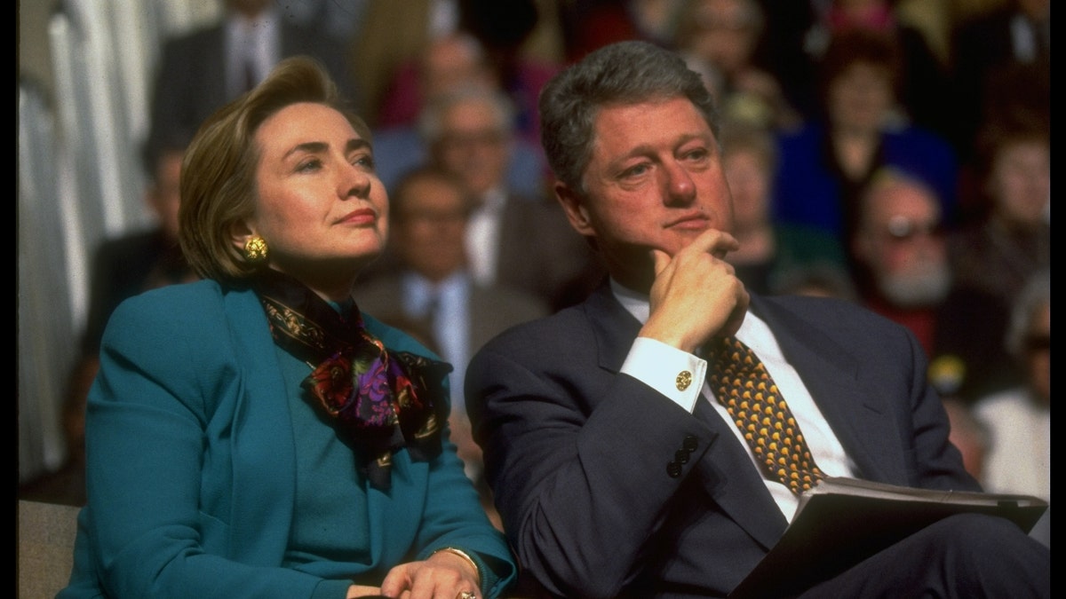 President Bill and Hillary Rodham Clinton sit together and listen to the speaker, surrounded by the audience at a health care event.