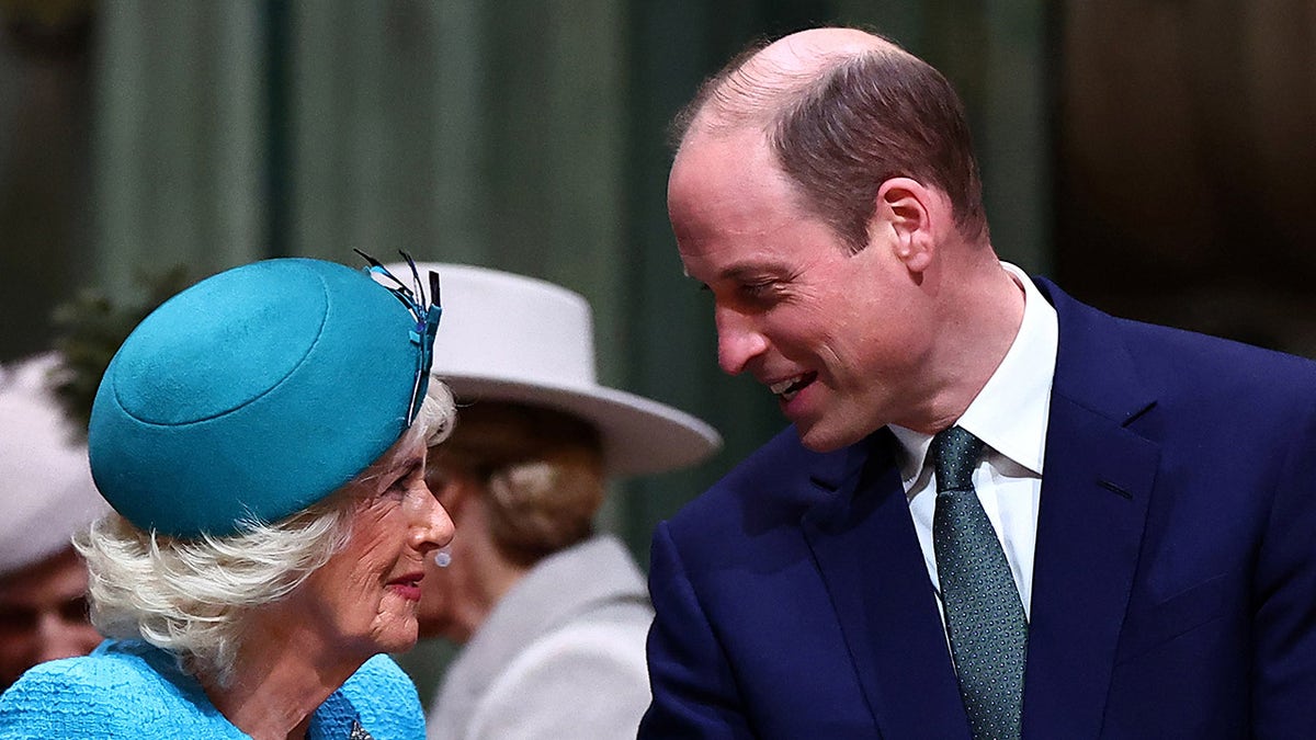 Queen Camilla wearing a blue dress and matching dress chatting with a smiling Prince William in a dark blue suit.