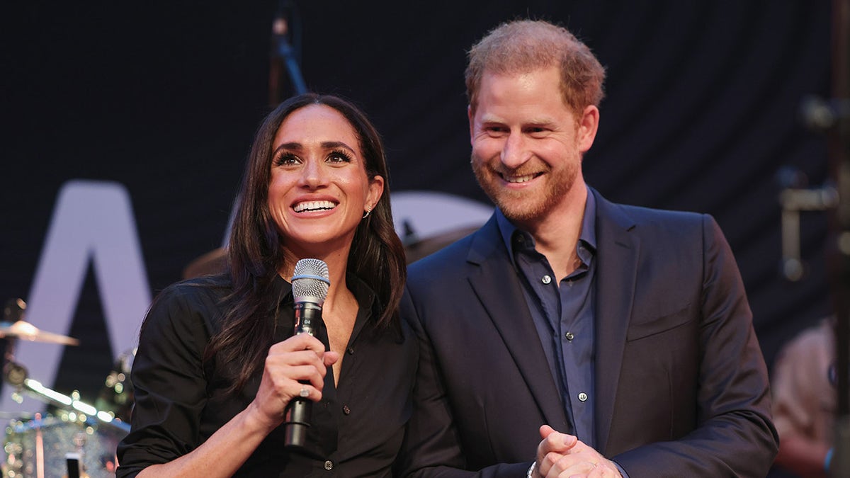 Meghan Markle con una camisa negra y un micrófono en la mano junto al príncipe Harry con una americana oscura y una camisa azul.