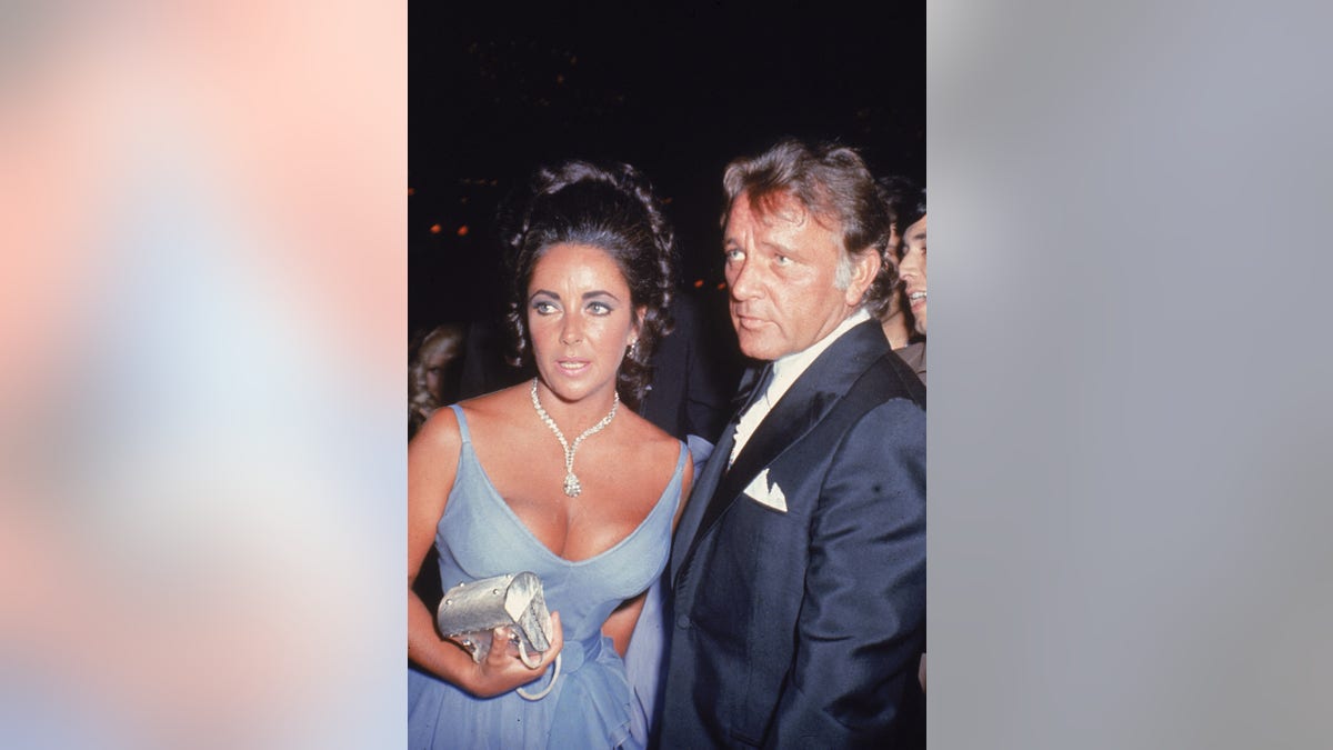 Elizabeth Taylor wearing a low cut lavender dress and a diamond necklace standing next to Richard Burton in a tux.