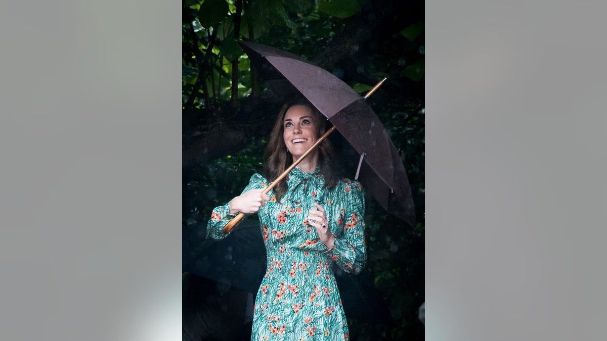 Kate Middleton con un vestido floral verde sonriendo y mirando hacia arriba y sujetando un paraguas.