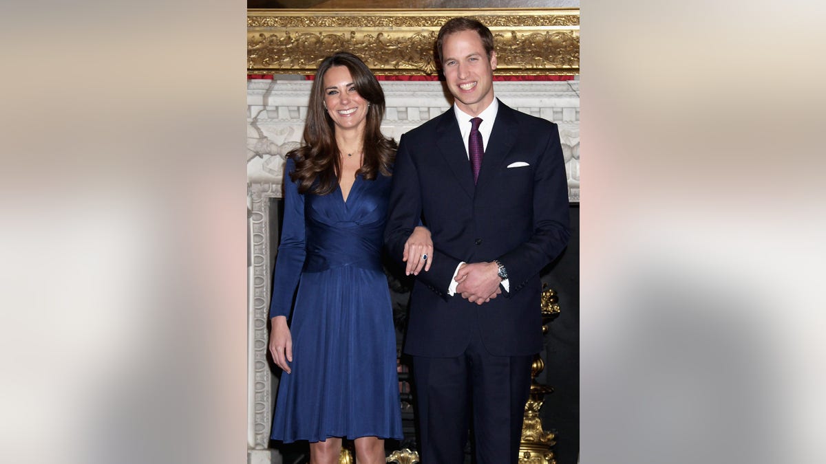 Kate Middleton wearing a royal blue gown showing off her engagement ring in the arm of Prince William wearing a suit.