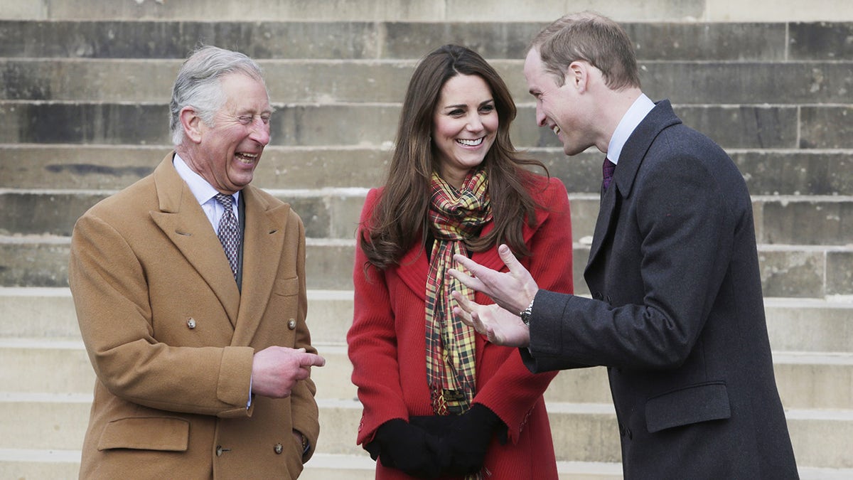 King Charles laughing at Prince William wearing a brown coat standing next to a smiling Kate Middleton wearing a red coat and a multiprinted scarf.