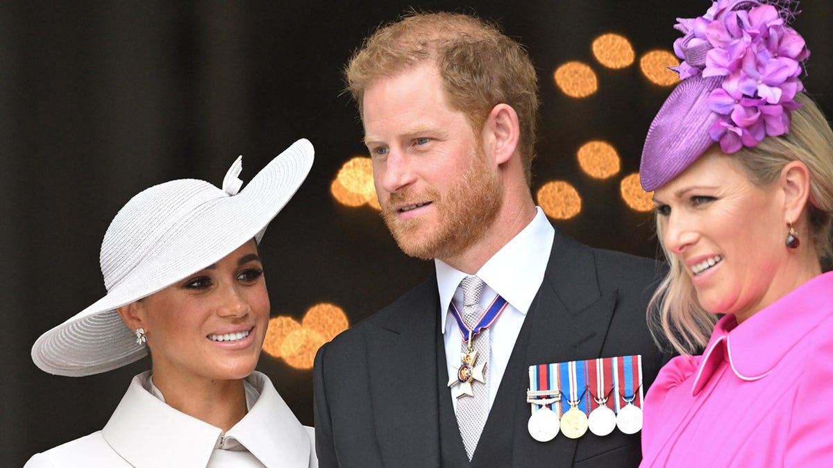 Meghan Markle in a white coat dress with a matching hat standing next to Prince Harry in a suit with medals and Zara Tindall wearing a pink coat dress with a matching fascinator.