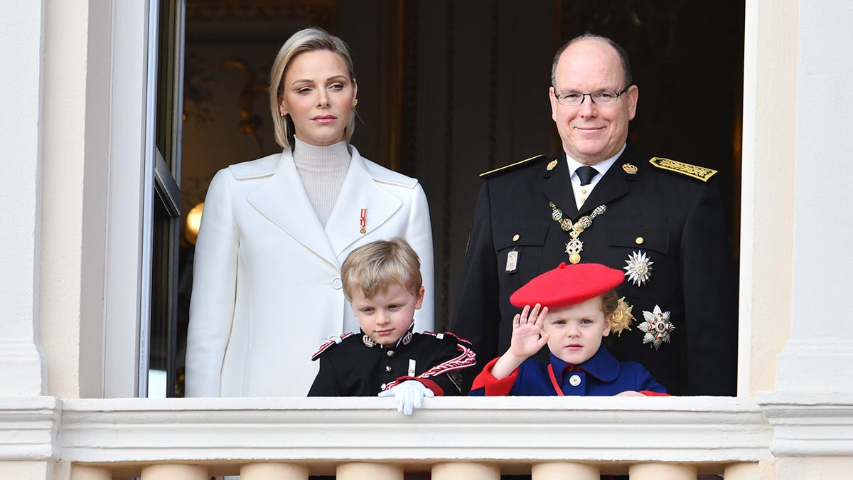 Princess Charlene wearing a white suit standing next to Prince Albert in a dark suit with medals and their two children.