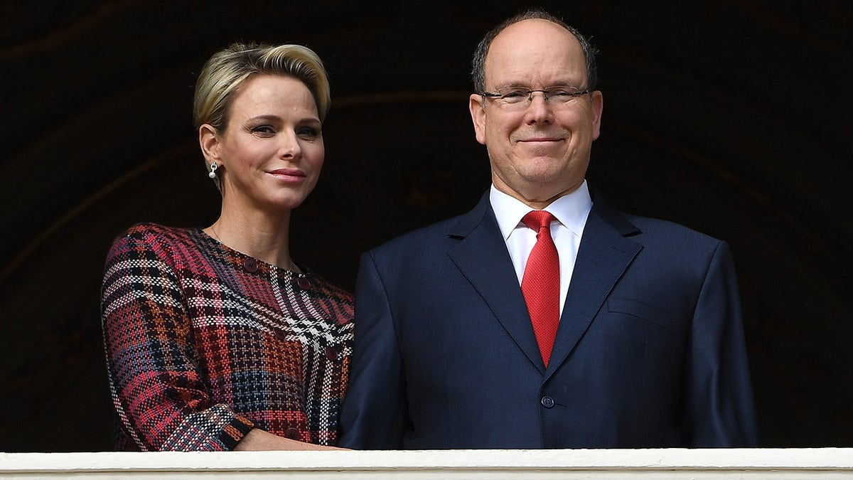 Princess Charlene of Monaco wearing a red and blue plaid dress standing next to Prince Albert in a navy suit and a red tie.