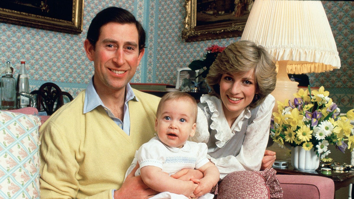 Prince Charles in a yellow sweater holding baby William next to a smiling Princess Diana inside their home.