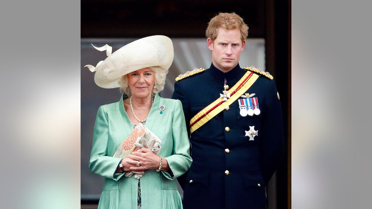 Queen Camilla wearing a green dress and a beige hat standing next to Prince Harry wearing a military uniform looking annoyed.