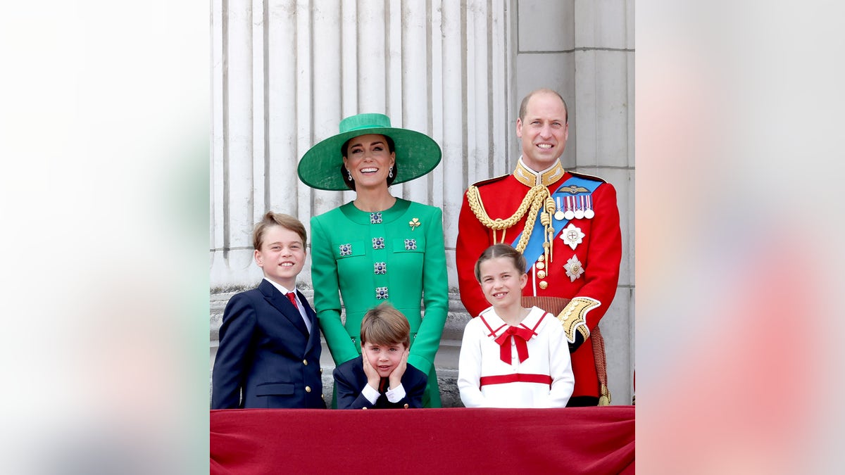 Prince William in a red uniform standing next to Kate Middleton in a green dress and matching hat on the balcony of Buckingham Palace with their three children.
