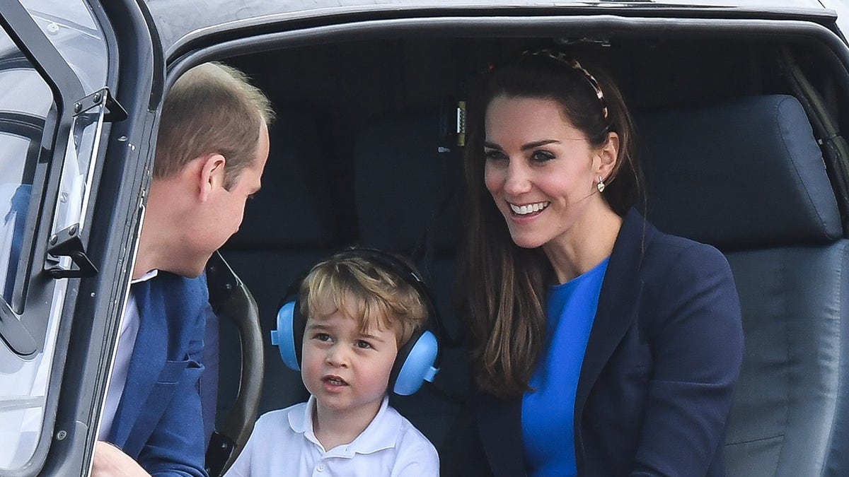 Prince William looking at Prince George wearing headphones as Kate Middleton smiles inside a helicopter.