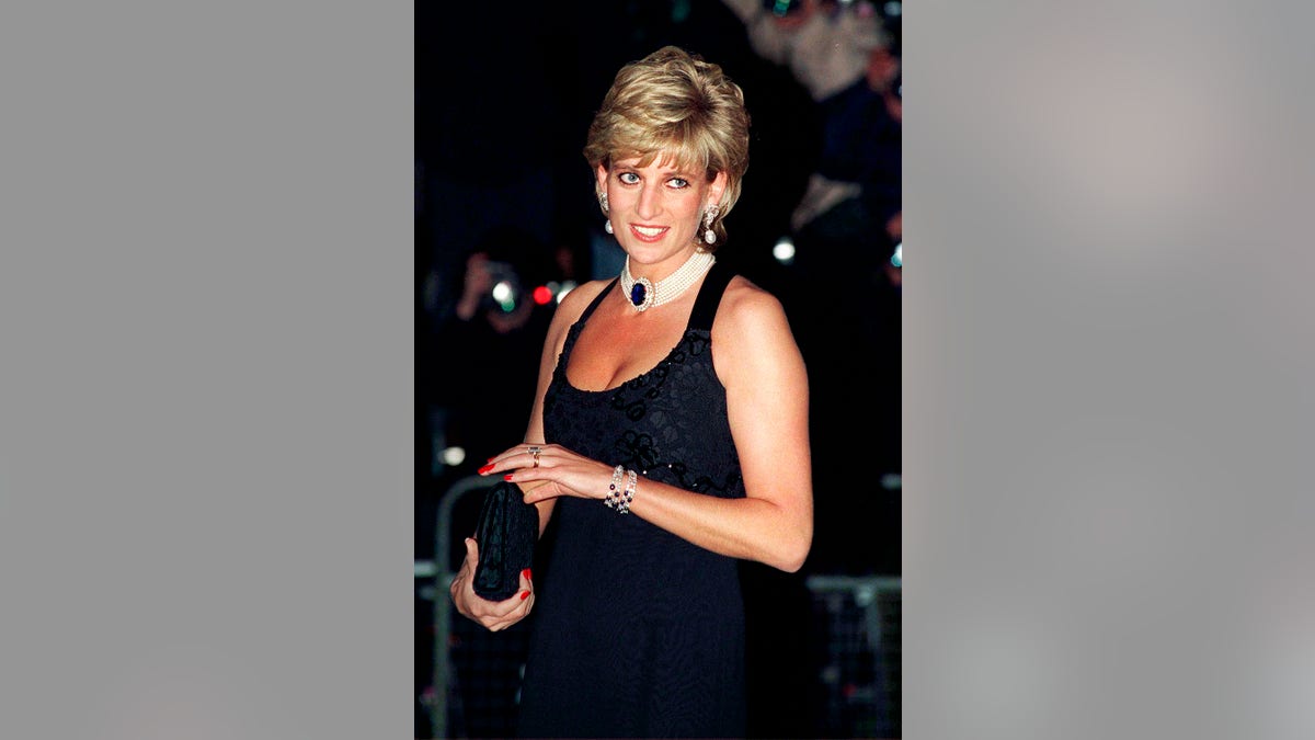 Princess Diana wearing a dark blue strappy dress with a pearl and sapphire choker as she holds a clutch.