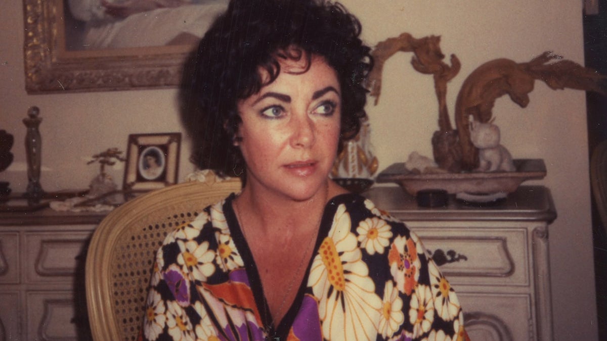A close-up of a candid photo of Elizabeth Taylor in a floral blouse looking away in a bedroom.