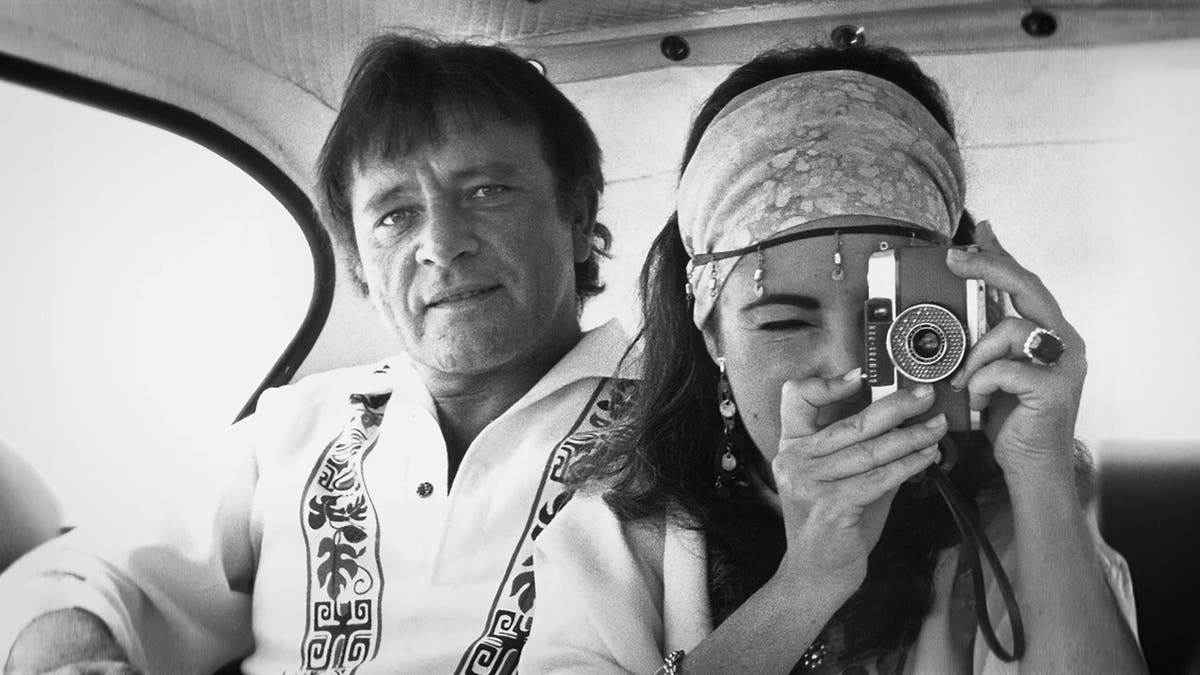 Elizabeth Taylor taking a photo while Richard Burton looks on from a car.