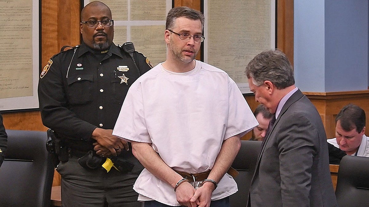 Shawn Grate in handcuffs in front of a police officer wearing a white shirt.