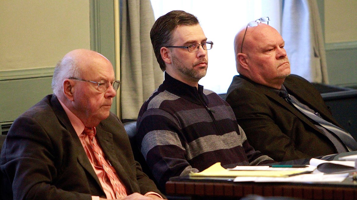 Shawn Grate wearing a striped grey sweater sitting in between two older men in court.