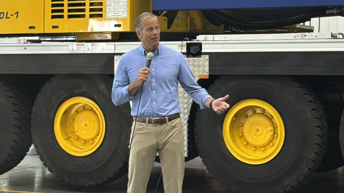 Senator John Thune memegang mikrofon di depan peralatan berat