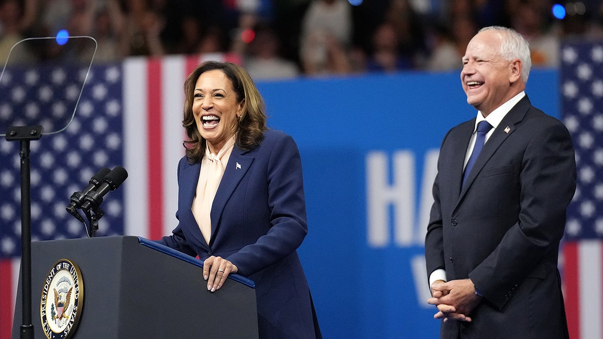 Kamala Harris tertawa di mimbar, Tim Walz di belakangnya di sebelah kirinya