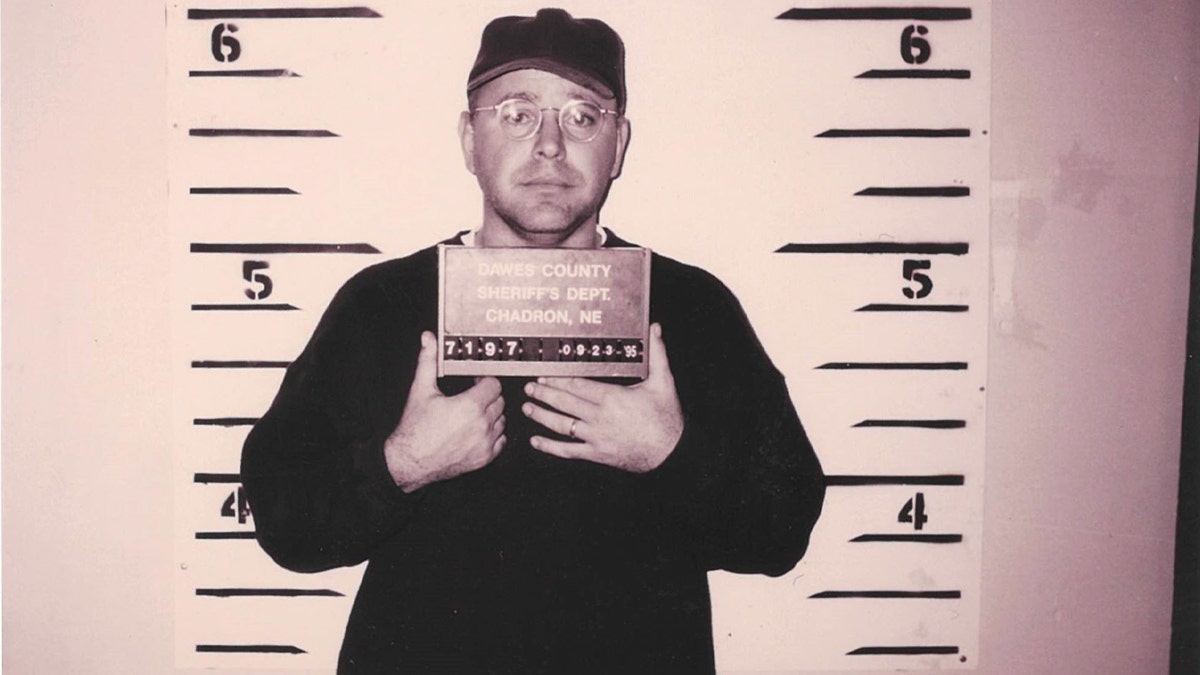 Tim Walz holding a plate and standing in front of a wall with height markers in a 1995 mugshot