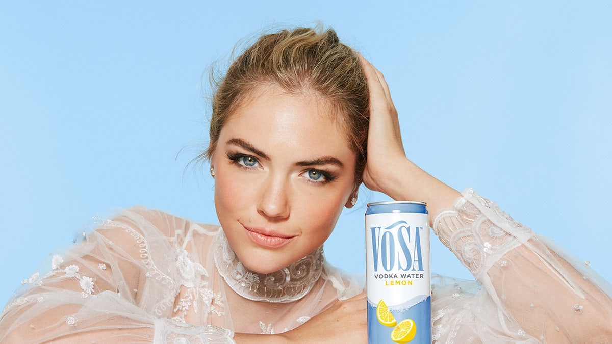 Kate Upton in a white high neck lace dress posing next to a can of Vosa Spirits