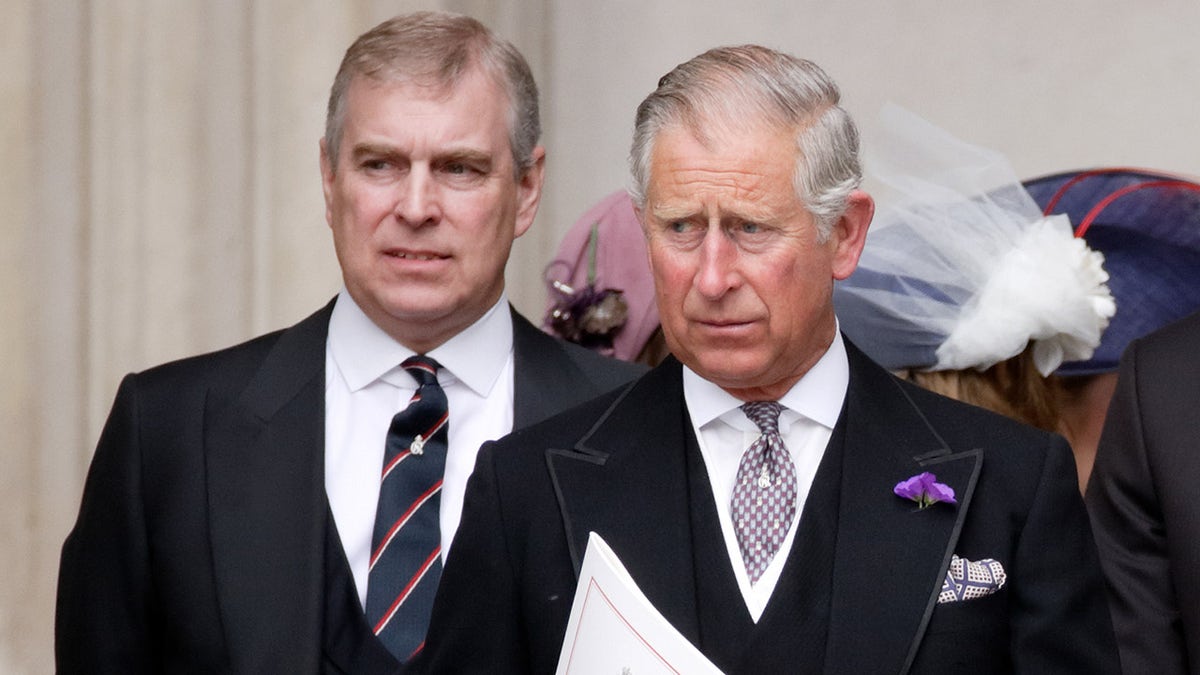 Prince Andrew in a black suit and striped tie looks tense walking behind his brother King Charles, also in a black suit and purple patterned tie