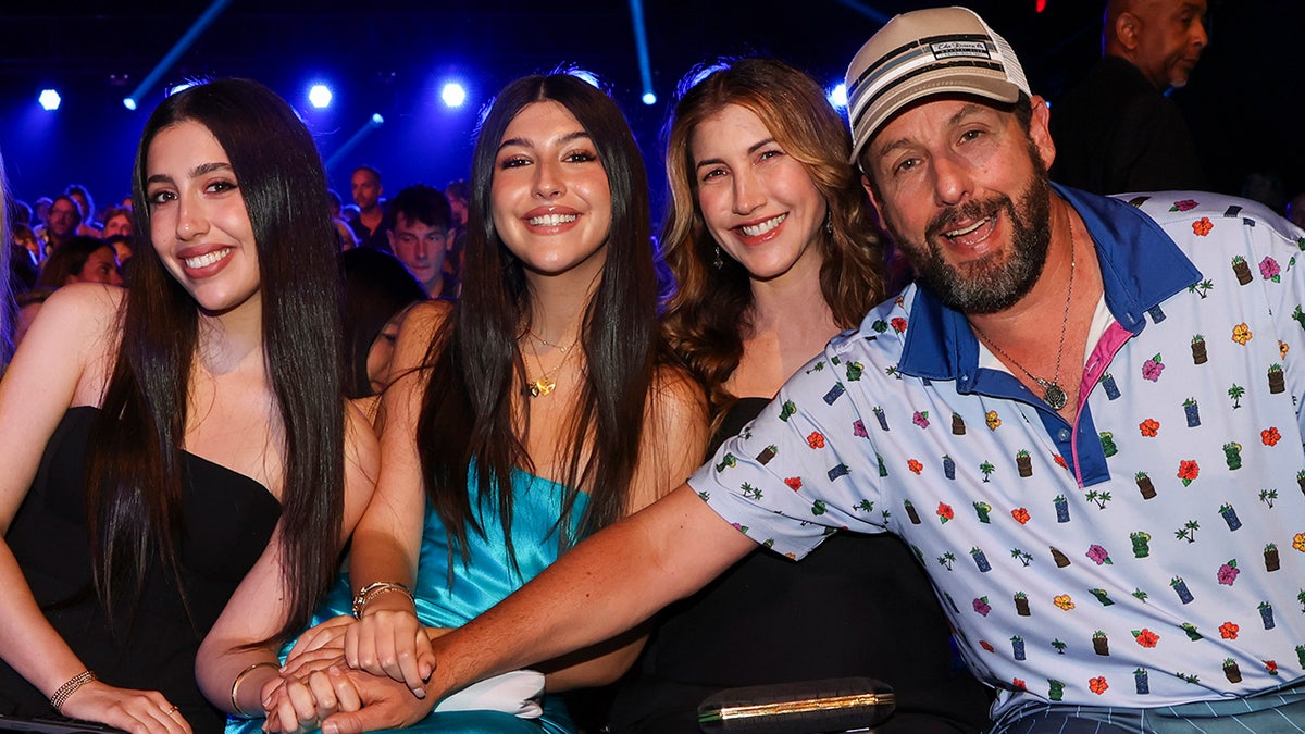 Adam Sandler in a blue collared shirt extends his arm over wife Jackie and reaches out to daughters Sadie and Sunny Sandler at the Kids Choice Awards