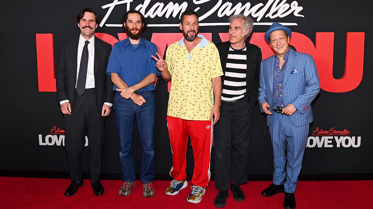 Dan Bulla in a black suit and tie smiles on the carpet next to Ronald Bronstein in a blue button down shirt and jeans next to Adam Sandler in a yellow patterned shirt next to Josh Safdie in a striped black and white shirt and black suit next to Rob Schneider in a matching blue suit and top hat