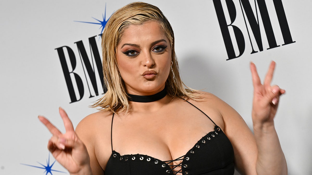 Bebe Rexha wearing a black necklace and a black corset-like shirt raises two peace signs on the carpet