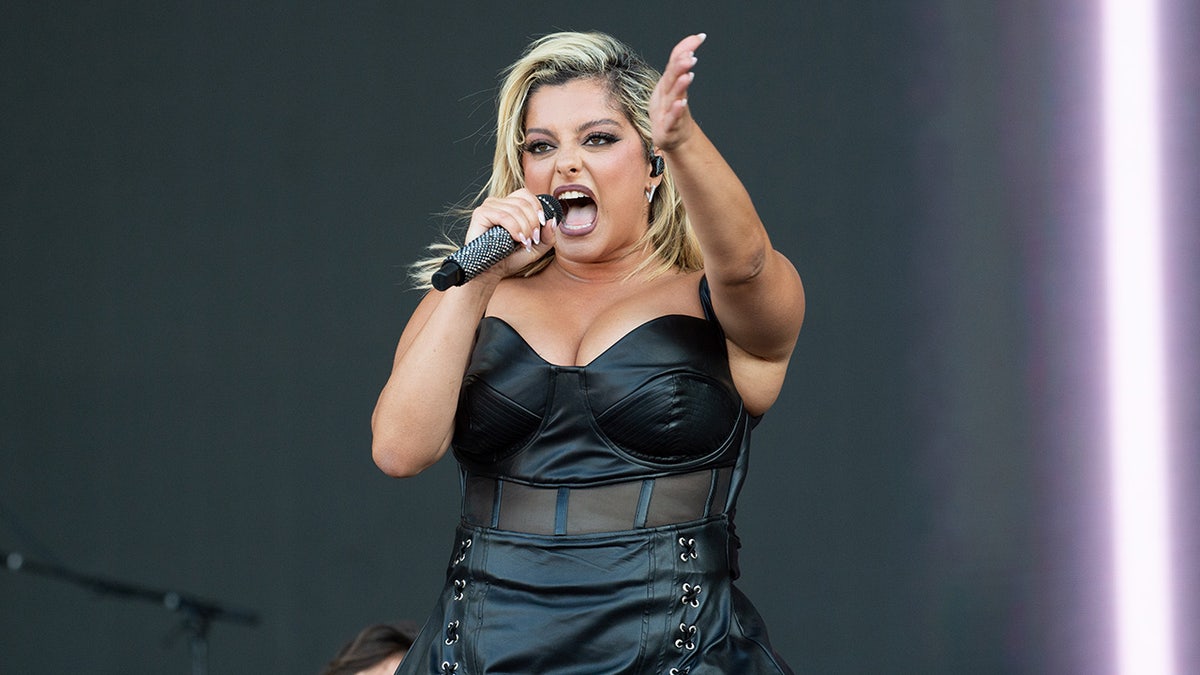 Bebe Rexha parece animada/zangada no palco cantando enquanto estende o braço em um espartilho de couro