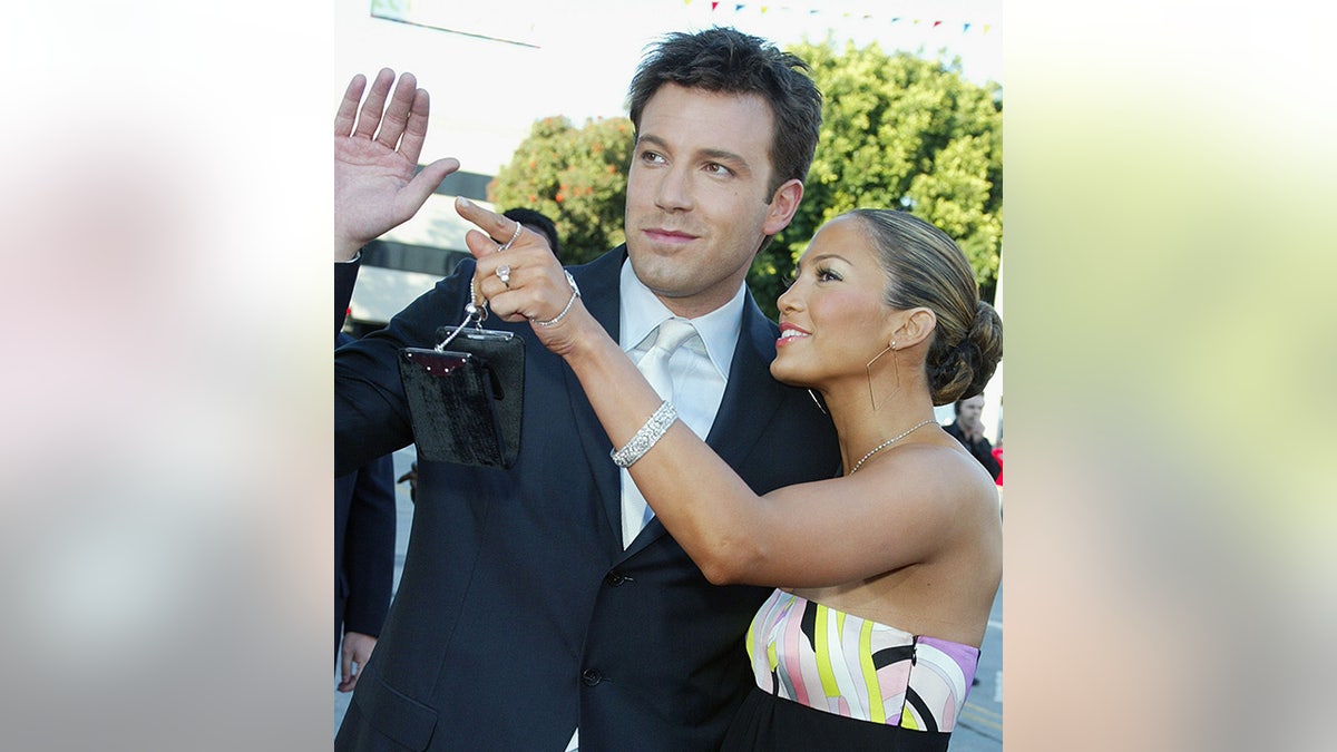 Ben Affleck waves as Jennifer Lopez in a colorful patterned strapless dress points towards something in the distance