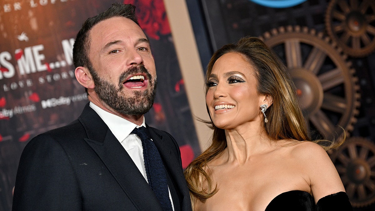 Ben Affleck em um terno preto e gravata está com a boca aberta no tapete enquanto Jennifer Lopez em um vestido preto olha para ele com adoração